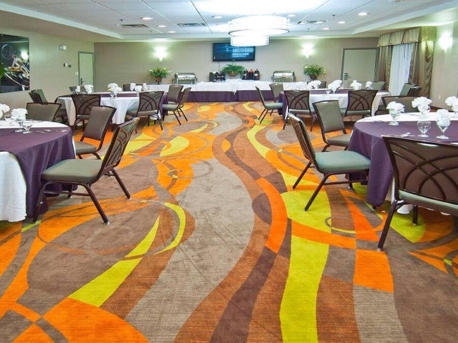 Un salón de conferencias con varias mesas y sillas, una alfombra estampada y una pantalla de televisión en la pared.