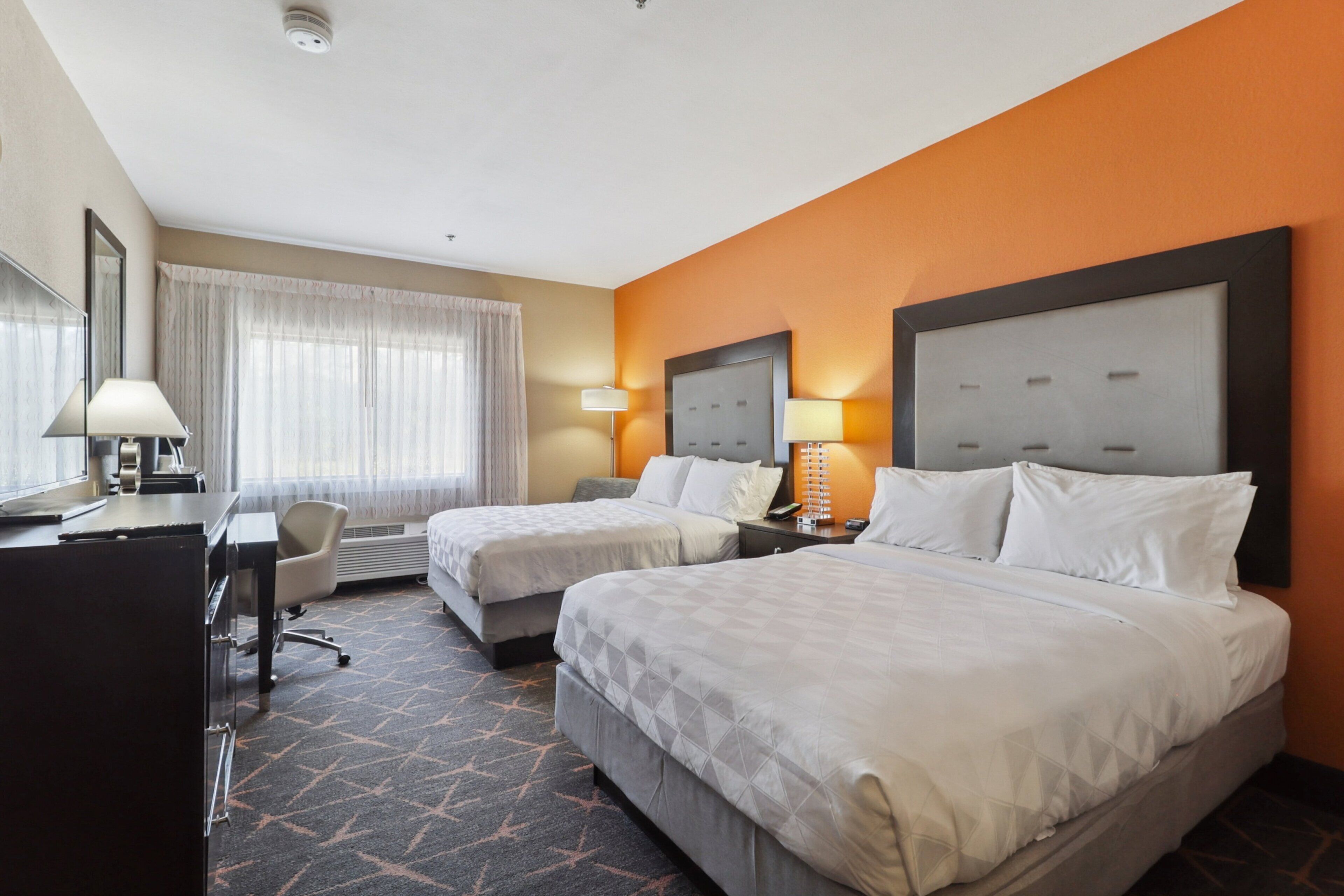 A hotel room with two beds, a desk, a chair, and a large window with curtains.