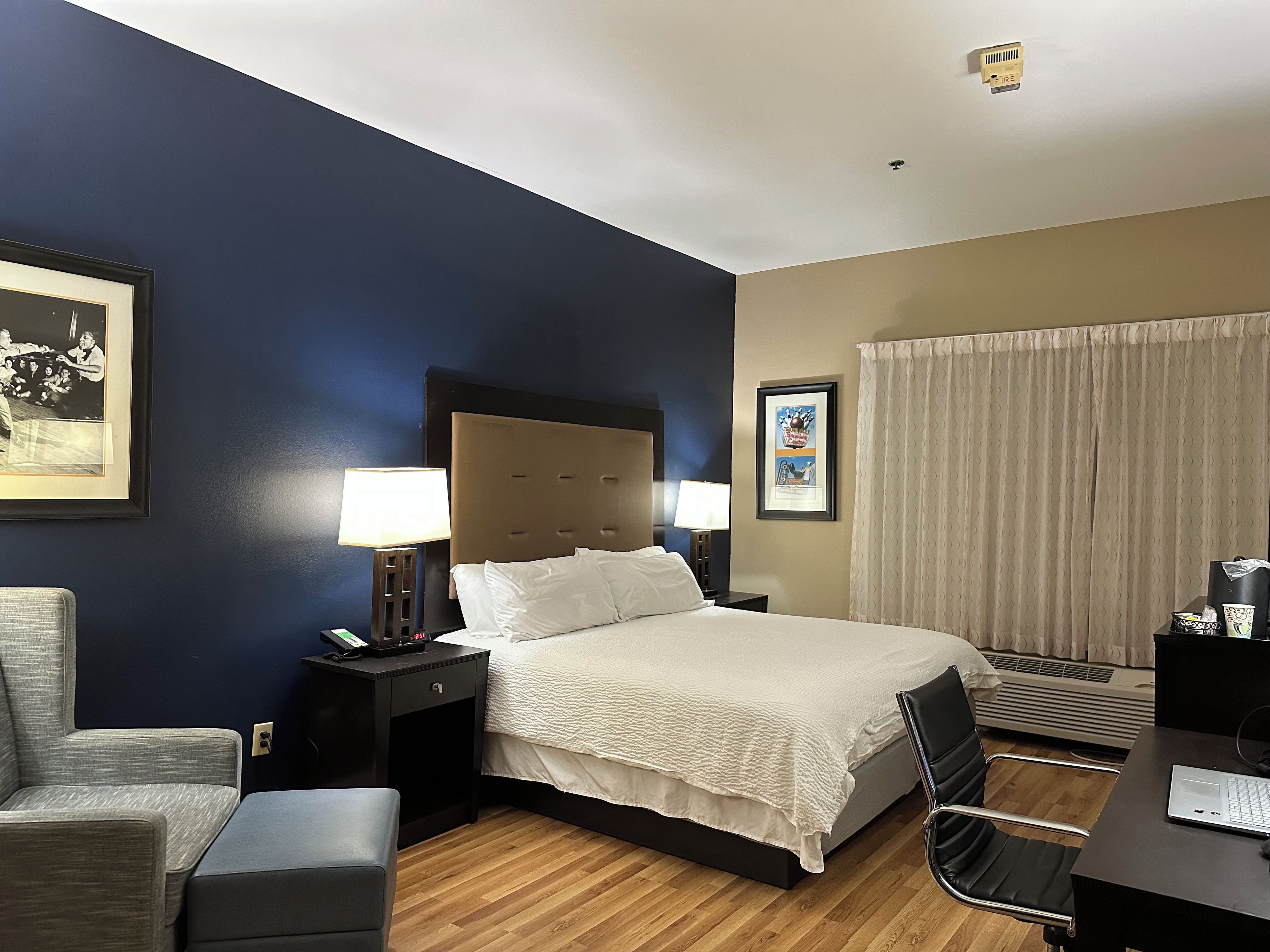 Una habitación de hotel con cama, mesitas de noche, escritorio, silla y ventana con cortinas.