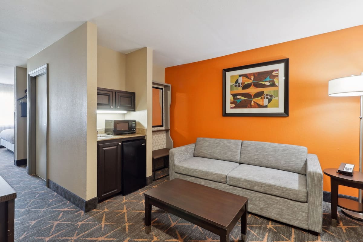 A hotel room with a kitchenette, a gray sofa, a coffee table, and a framed abstract painting on the orange wall.