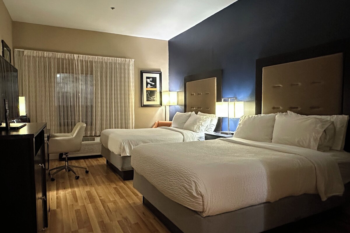 Habitación de hotel con dos camas, un escritorio, una silla y un amplio ventanal con cortinas.