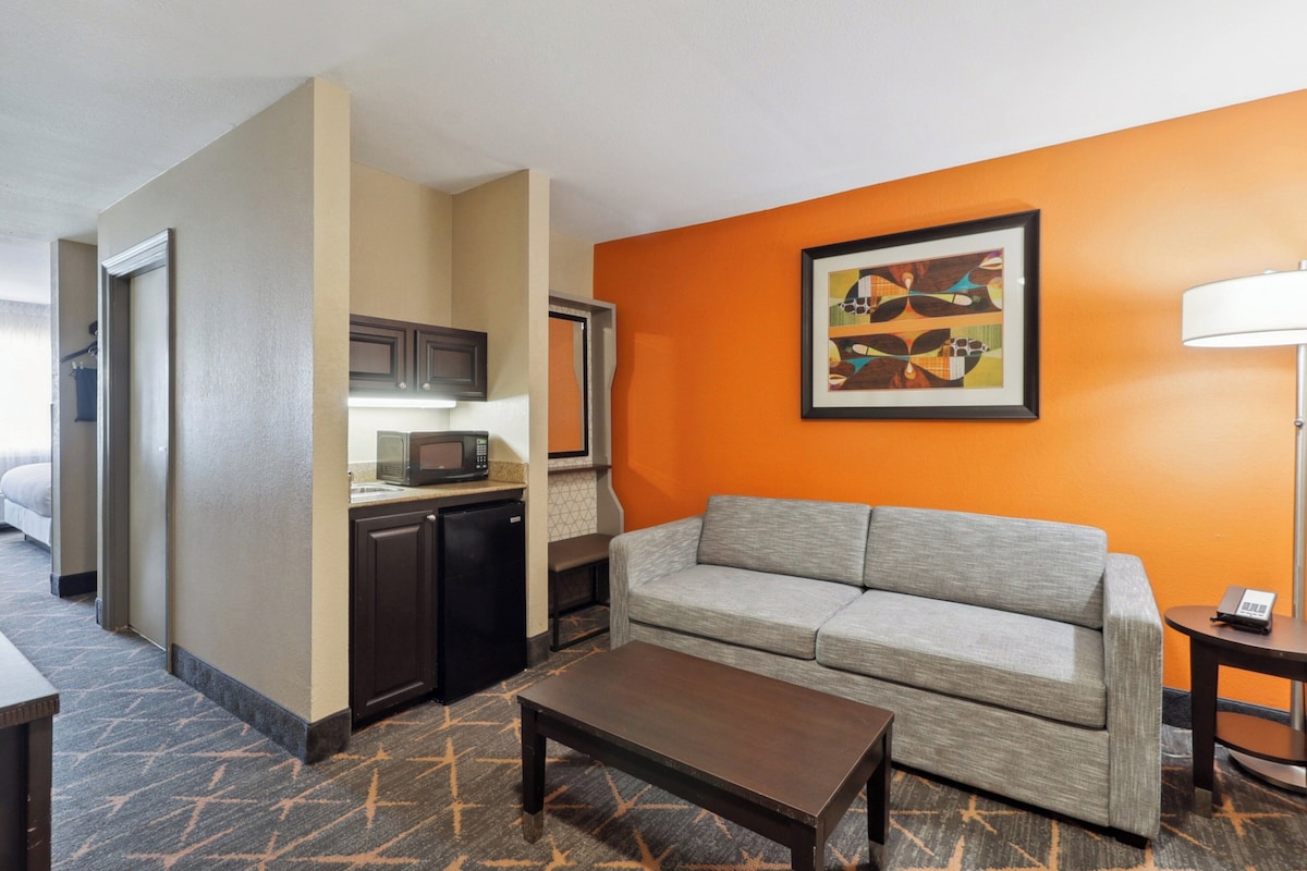 A hotel room with a kitchenette, a gray sofa, a coffee table, and a framed abstract painting on the orange wall.