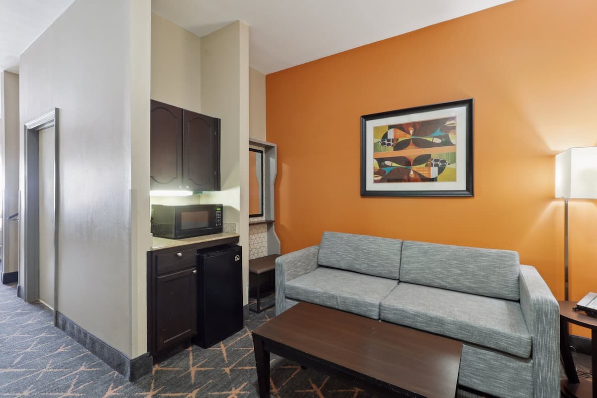 A hotel room with a kitchenette, a gray sofa, a wooden coffee table, and a framed abstract painting on the orange wall.