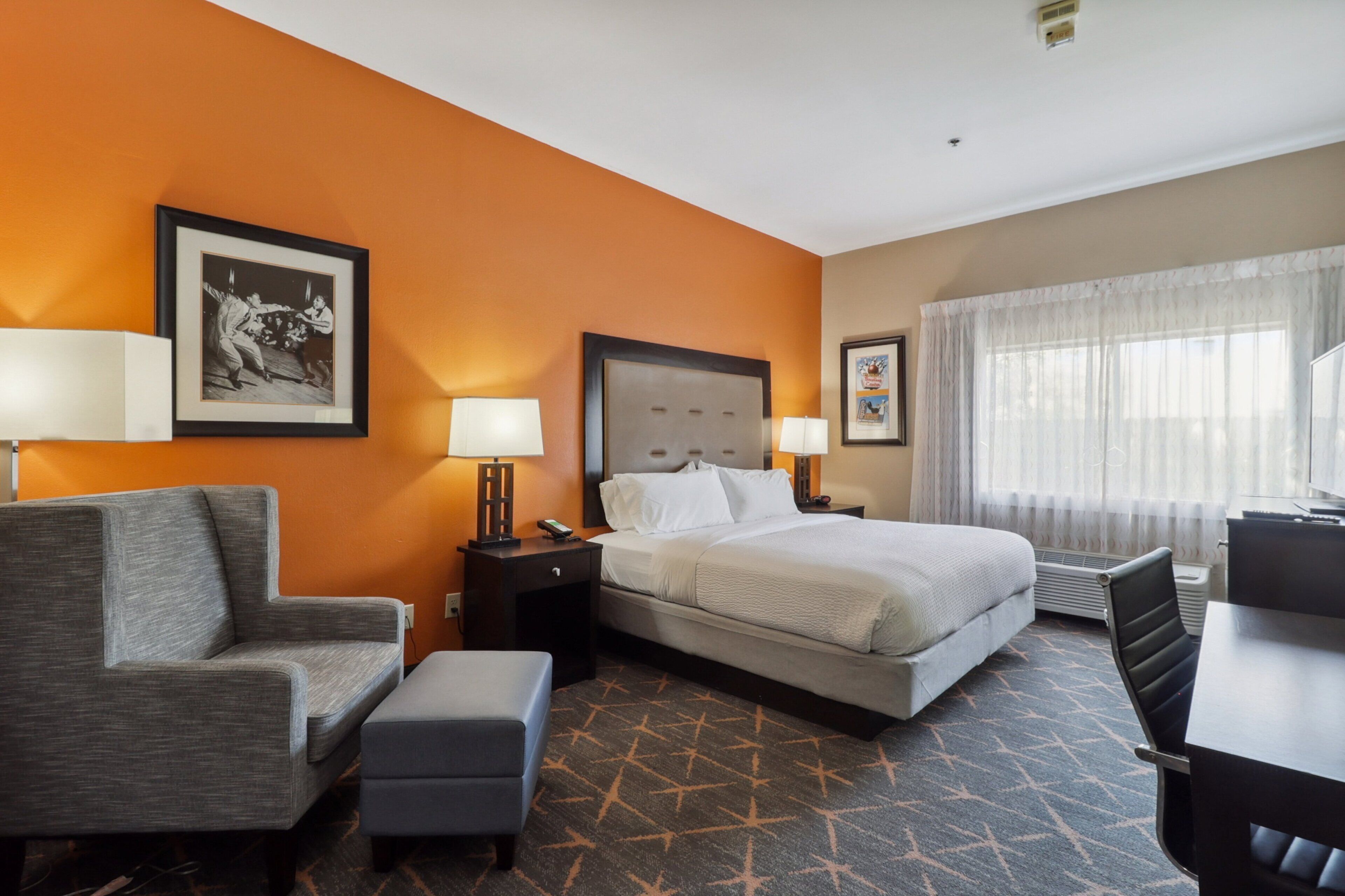 A hotel room with a bed, a desk, a chair, a nightstand, and a lamp.