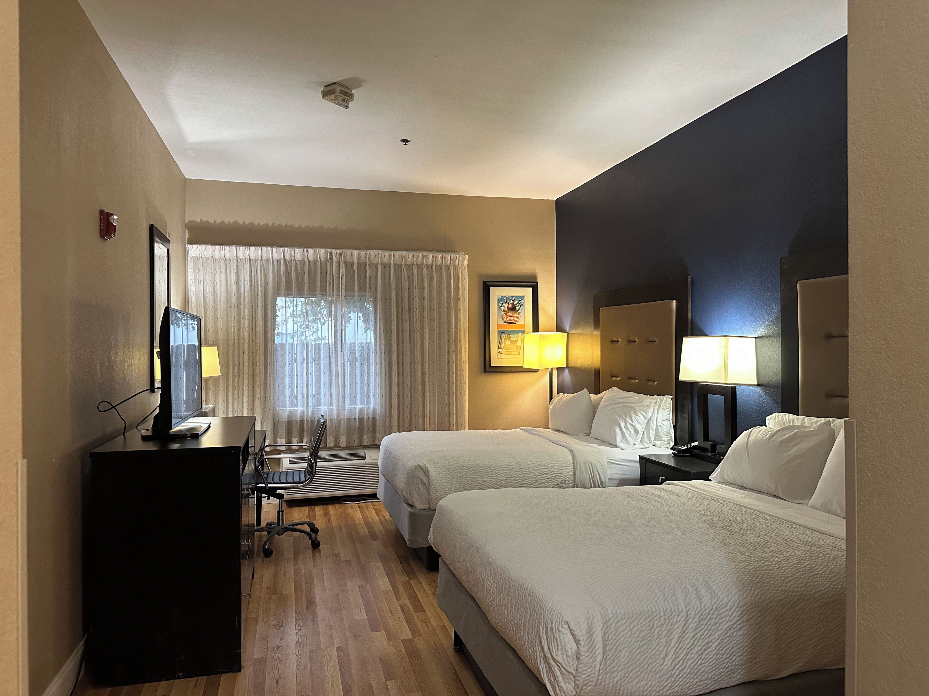 A hotel room with two beds, a desk with a computer, a window with curtains, and a ceiling fan.