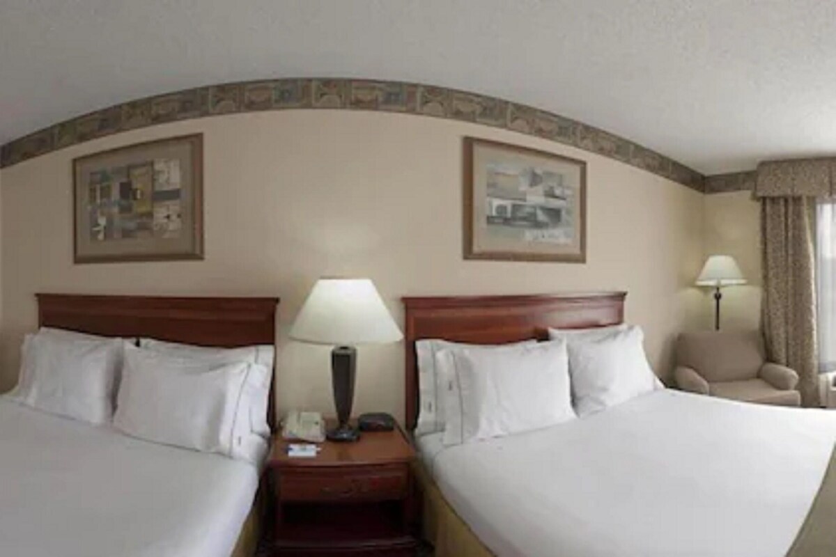 A hotel room with two beds, a nightstand with a lamp, and a chair.