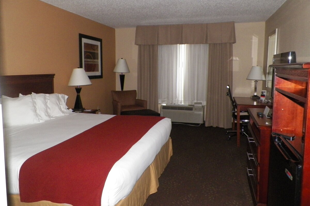 A hotel room with a large bed, two bedside tables with lamps, a desk with a chair, a dresser, and a window with curtains.