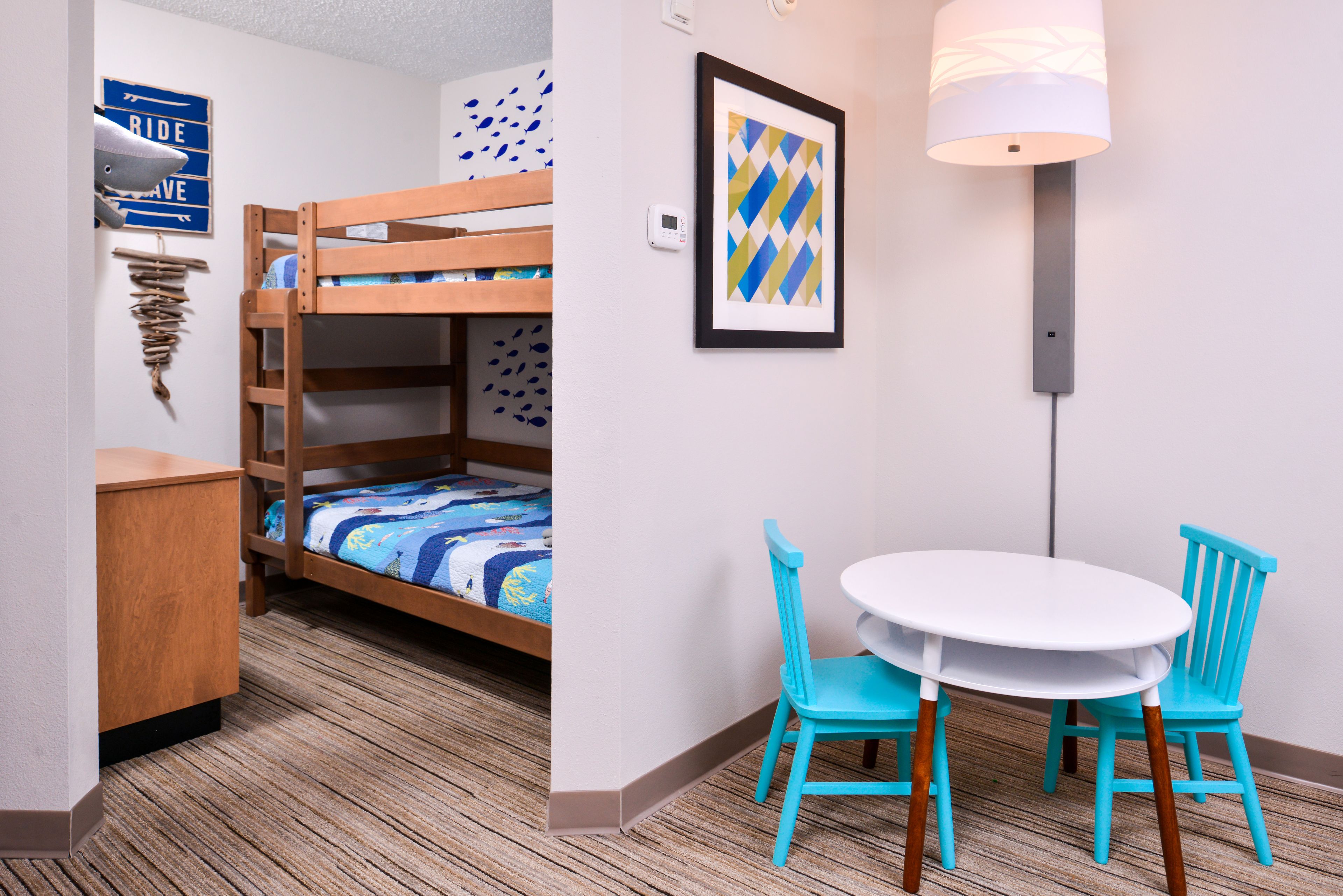Una cama litera con colcha azul y blanca a rayas, un escritorio de madera pequeño, un cuadro en la pared y una mesa redonda con dos sillas turquesas.