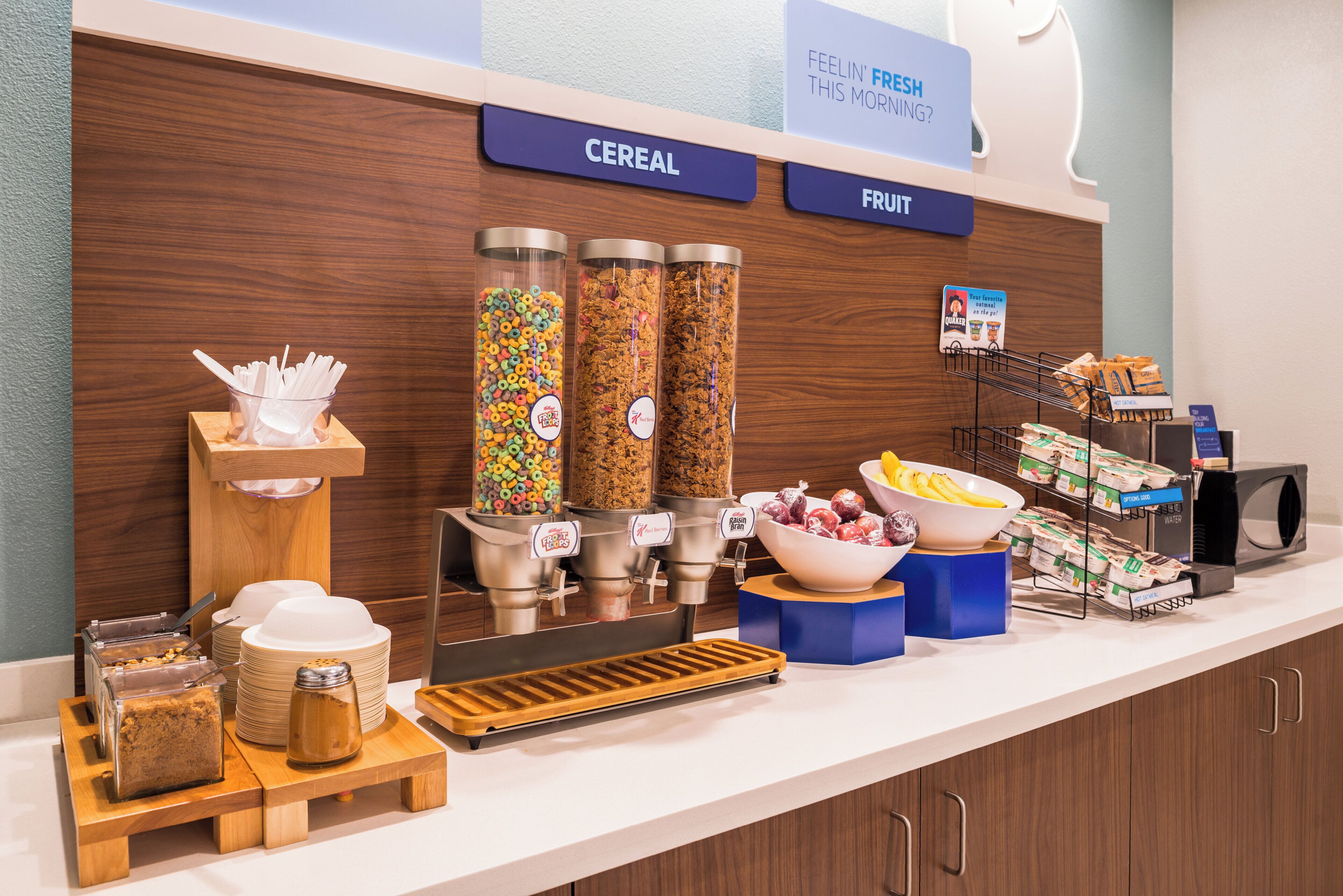 A breakfast buffet with cereal and fruit options, a coffee machine, and various breakfast items.