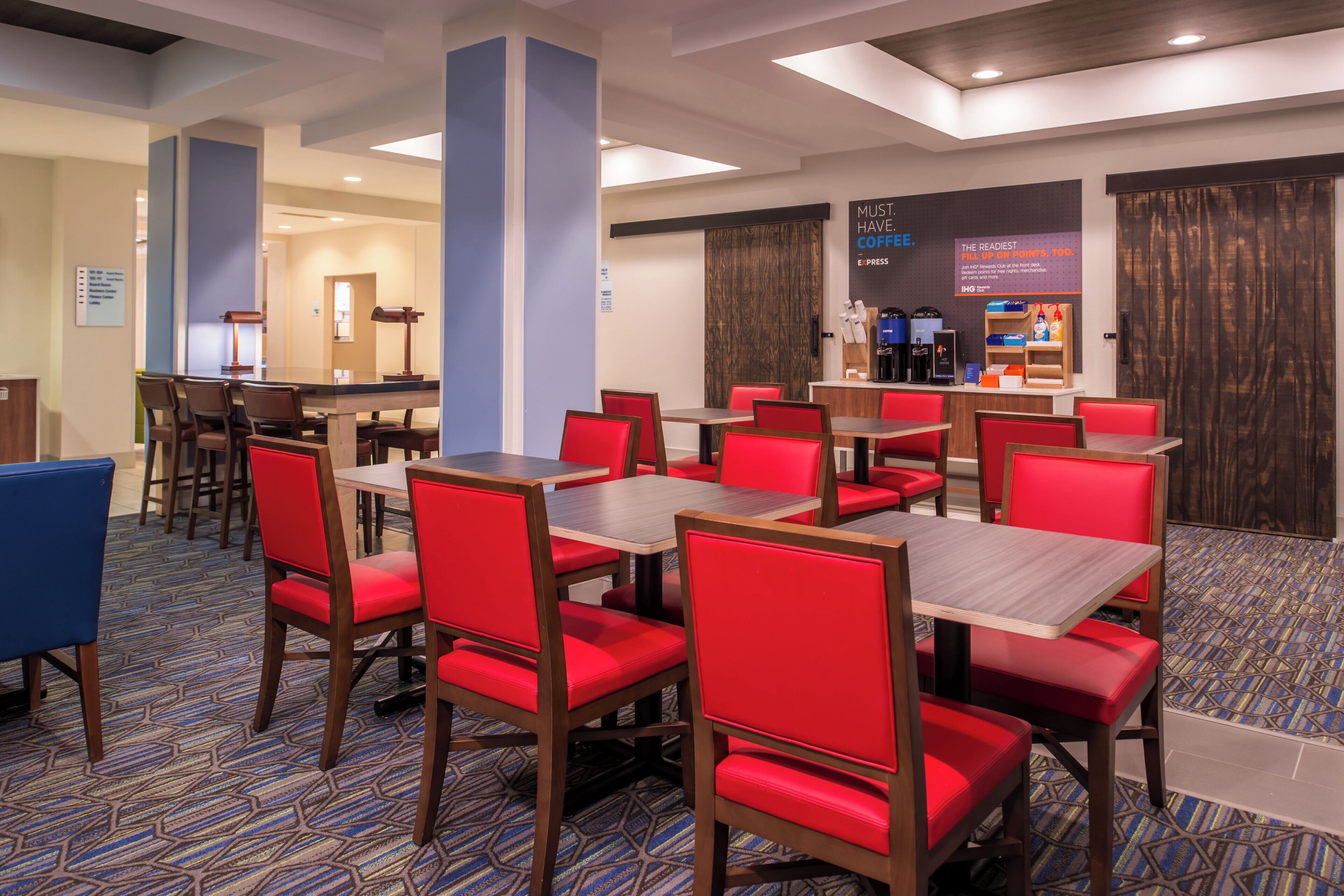 A hotel dining area with red upholstered chairs and wooden tables, a coffee station, and a sign indicating a rewards program.