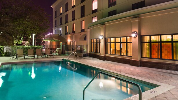 A swimming pool with a clear edge, surrounded by a paved deck and a building with multiple windows.