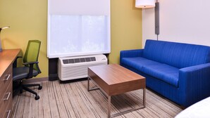 A hotel room with a blue sofa, a wooden table, a green office chair, a projector screen, and a wall-mounted lamp.
