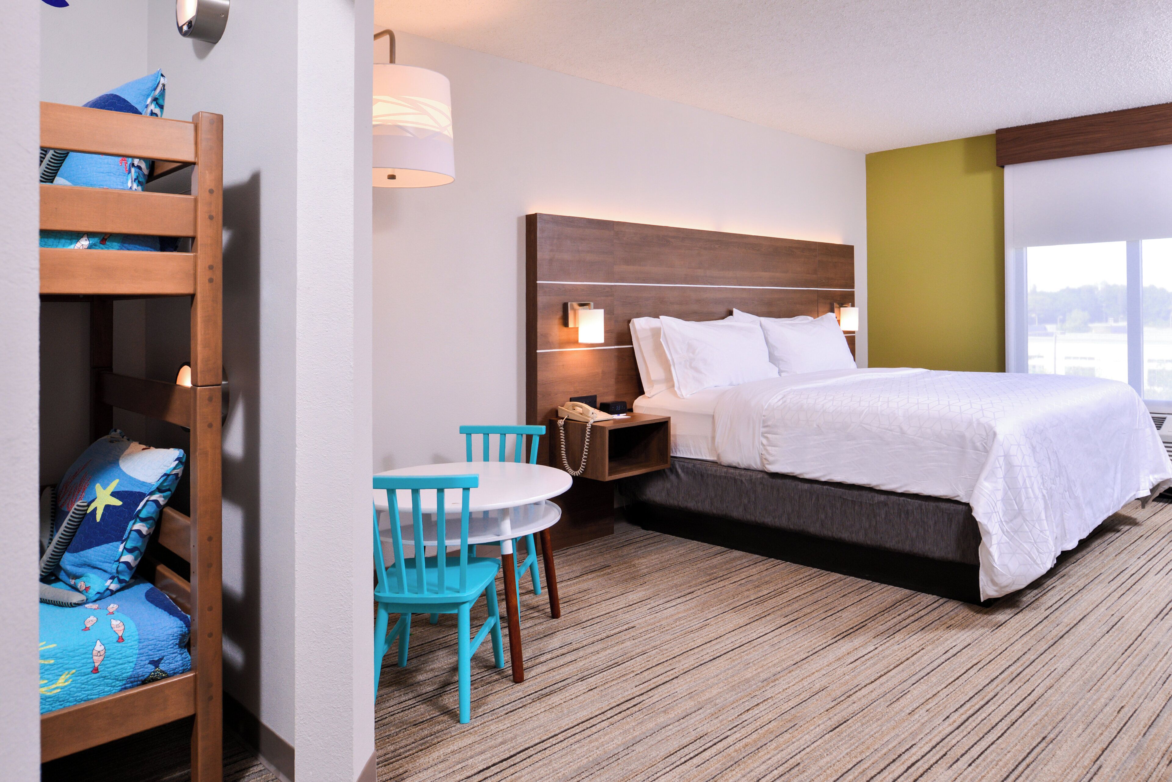 A hotel room with a bunk bed, a single bed, a small table with chairs, a nightstand, and a lamp.