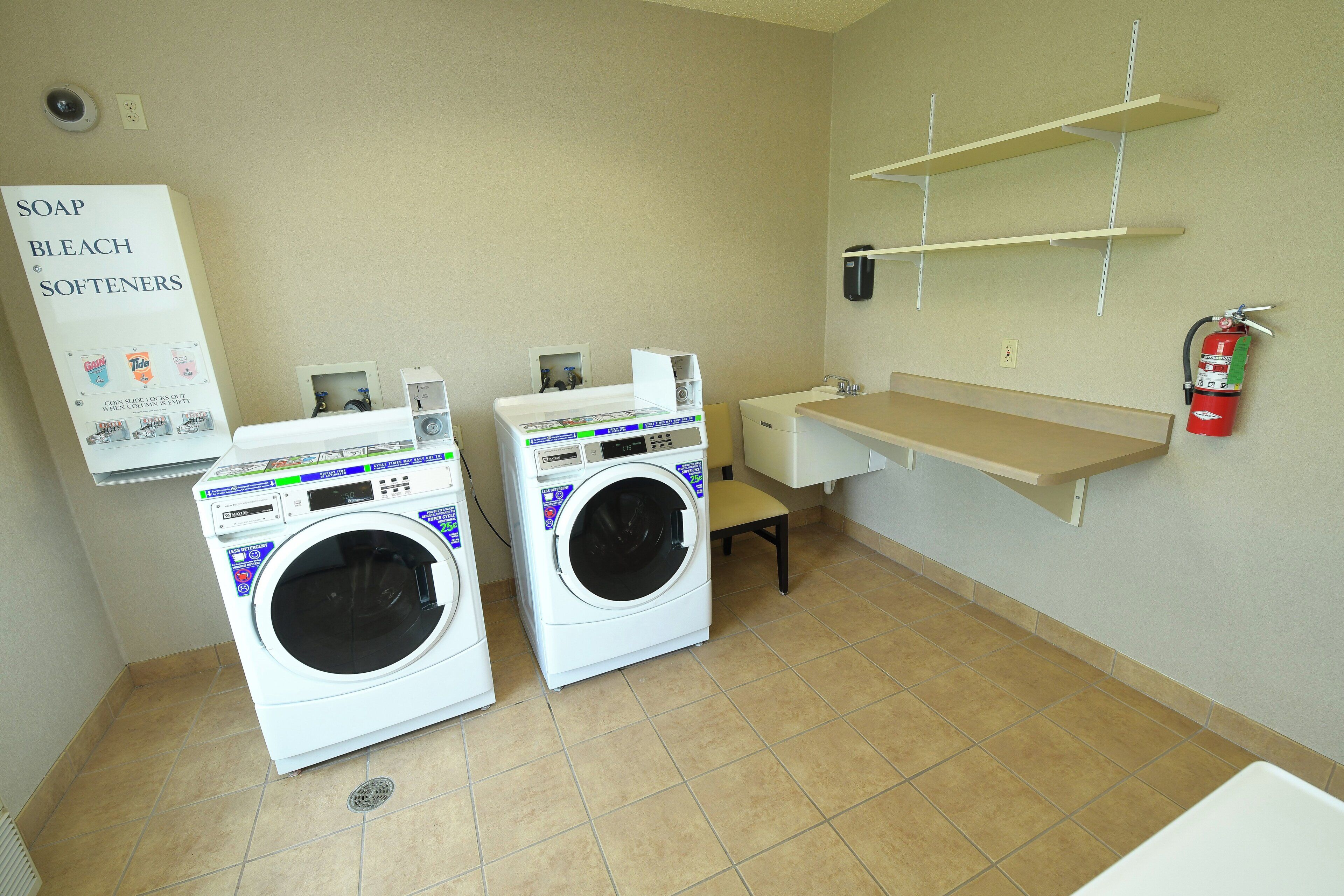 A laundry room with two front-loading washing machines, a shelf with a fire extinguisher, and a sign for soap and bleach softeners.