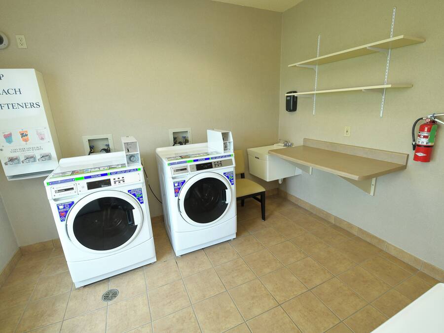A laundry room with two front-loading washing machines, a shelf with a fire extinguisher, and a sign for soap and bleach softeners.