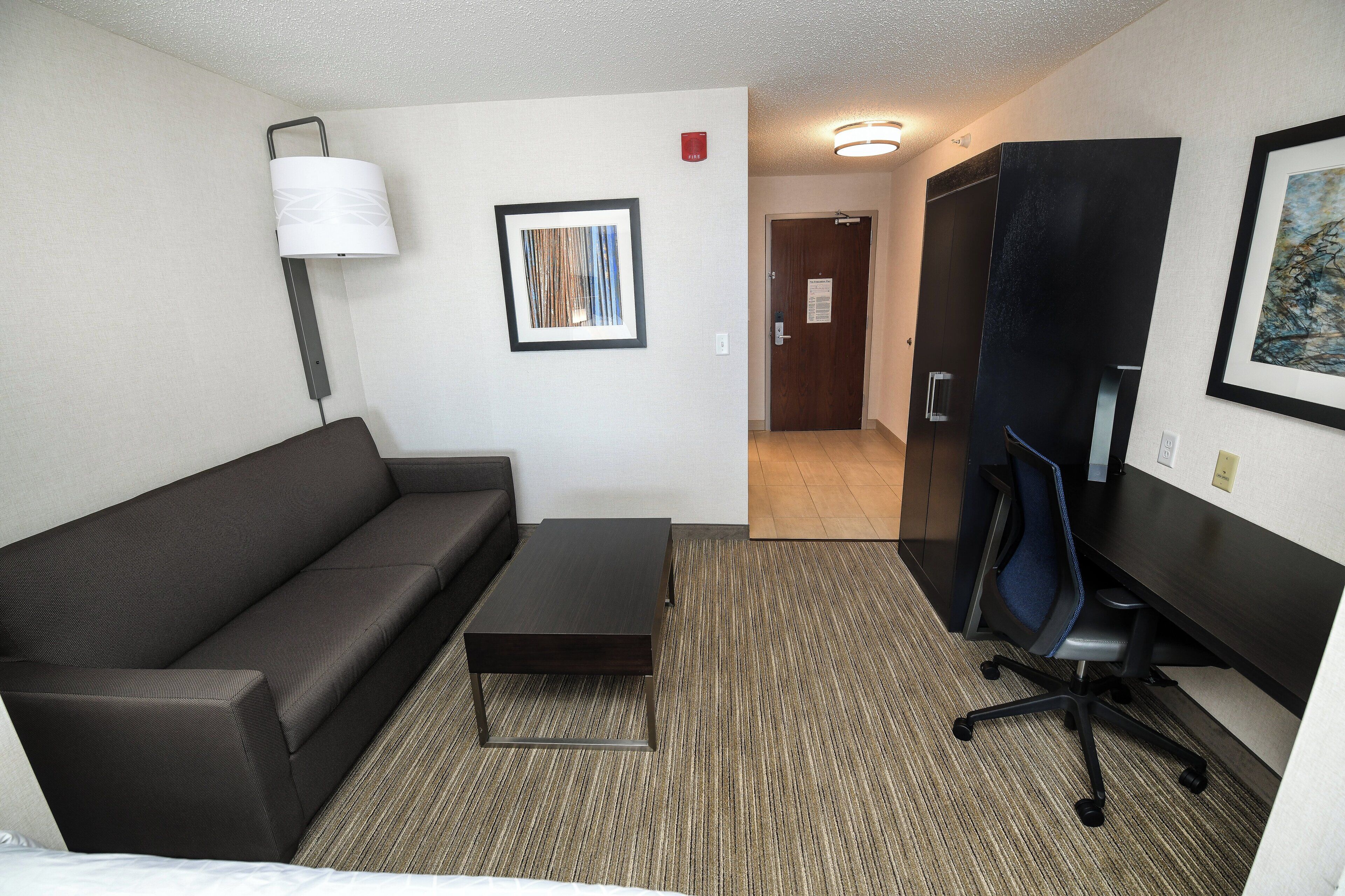 Una habitación de hotel con un sofá marrón oscuro, una pequeña mesa de centro, un escritorio con una silla, una lámpara, un cuadro abstracto enmarcado y una puerta que da a otra habitación.