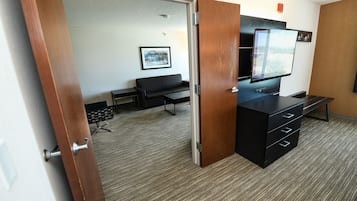 A hotel room with a wooden door, a black sofa, a flat-screen TV, and a desk with drawers.