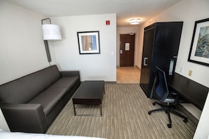 Une chambre d’hôtel avec un canapé marron foncé, une petite table basse en bois, une seule œuvre d’art encadrée au mur, un lampadaire, un bureau avec une chaise et une porte donnant sur une autre pièce.