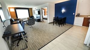 Habitación de hotel moderna con una cama grande, un escritorio con silla, una zona de comedor con sillas, un área de cocina con lavavajillas y una pared de acento azul.