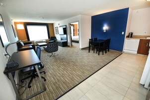 Habitación de hotel moderna con una cama grande, un escritorio con silla, una zona de comedor con sillas, un área de cocina con lavavajillas y una pared de acento azul.