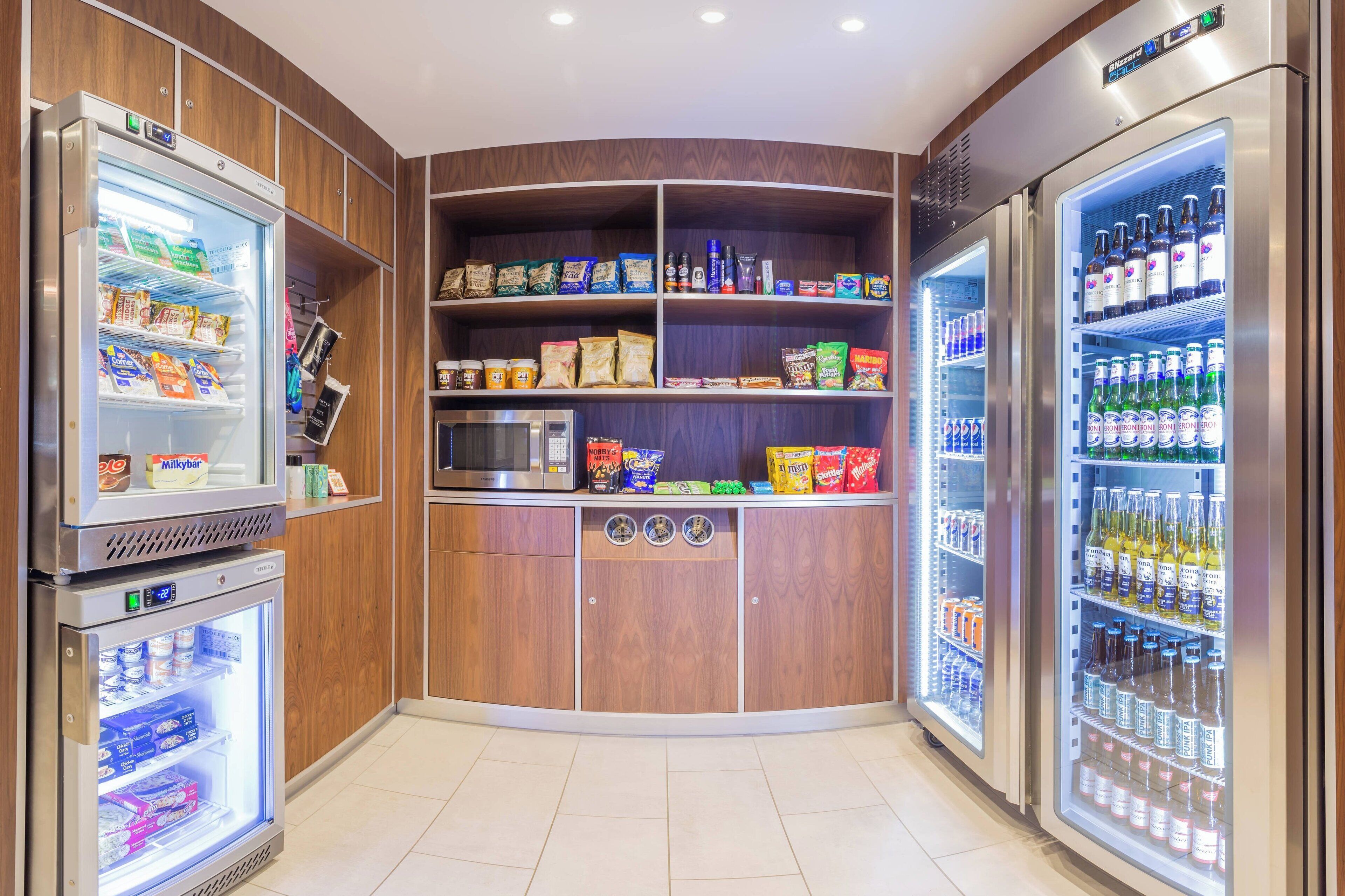 Una kitchenette en esquina con microondas, refrigerador y congelador. Estantes llenos de bocadillos, bebidas y condimentos.