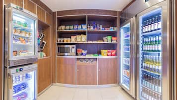 A corner kitchenette with a microwave, a refrigerator, and a freezer. Shelves are stocked with snacks, beverages, and condiments.