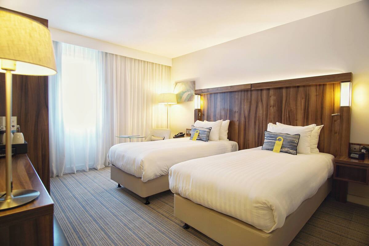 Ein Hotelzimmer mit zwei Betten, einem Schreibtisch, einer Lampe und einem Nachttisch.