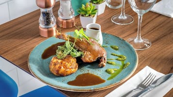 A plate of food with a roasted chicken leg, a piece of meat, and a sauce, accompanied by a small cup of sauce and a green sauce on a blue plate.