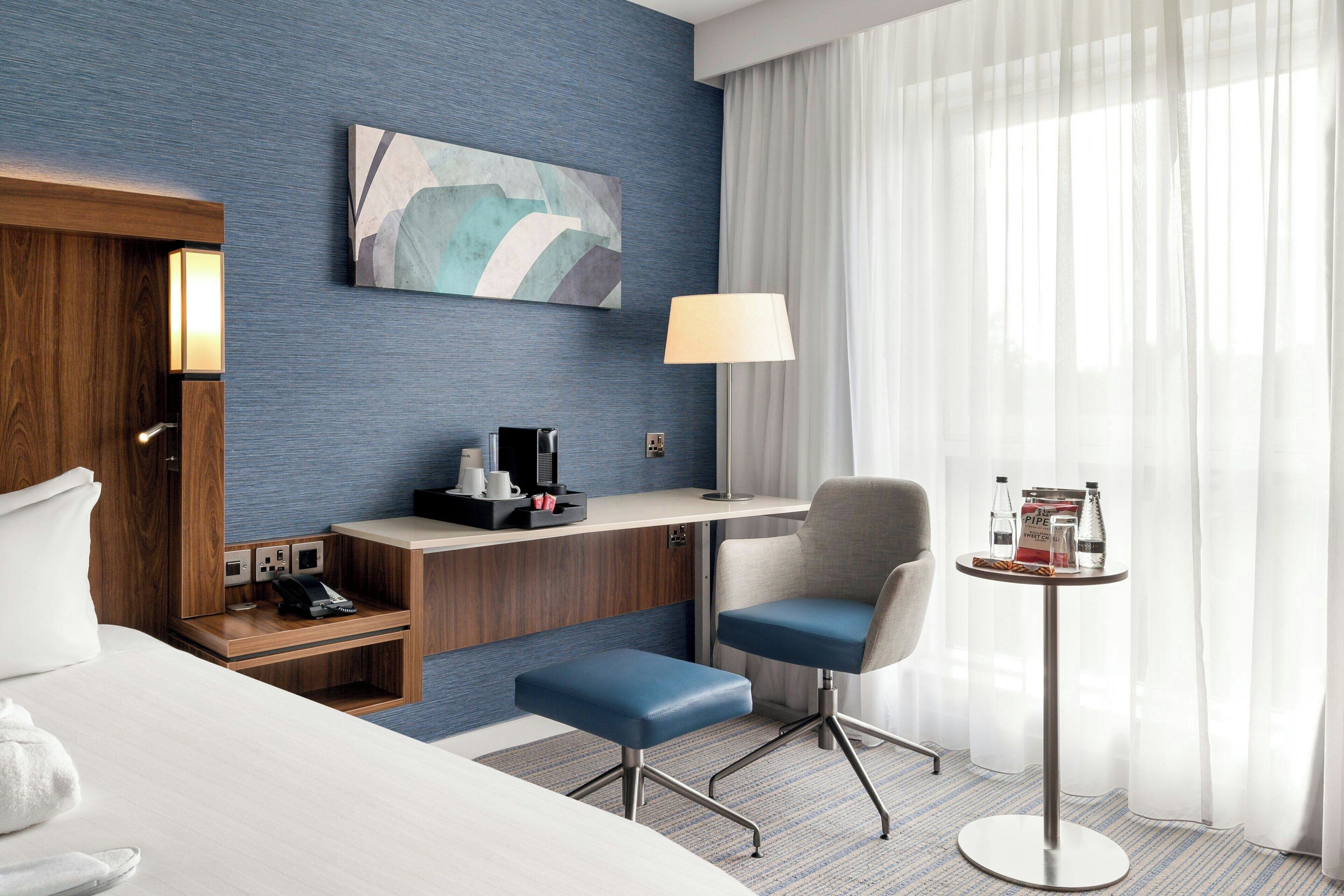 A hotel room with a bed, a desk, a chair, a small table, a lamp, and a painting on the wall.