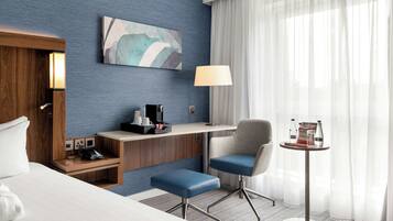 A hotel room with a bed, a desk, a chair, a small table, a lamp, and a painting on the wall.
