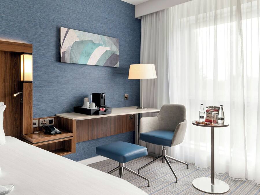A hotel room with a bed, a desk, a chair, a small table, a lamp, and a painting on the wall.