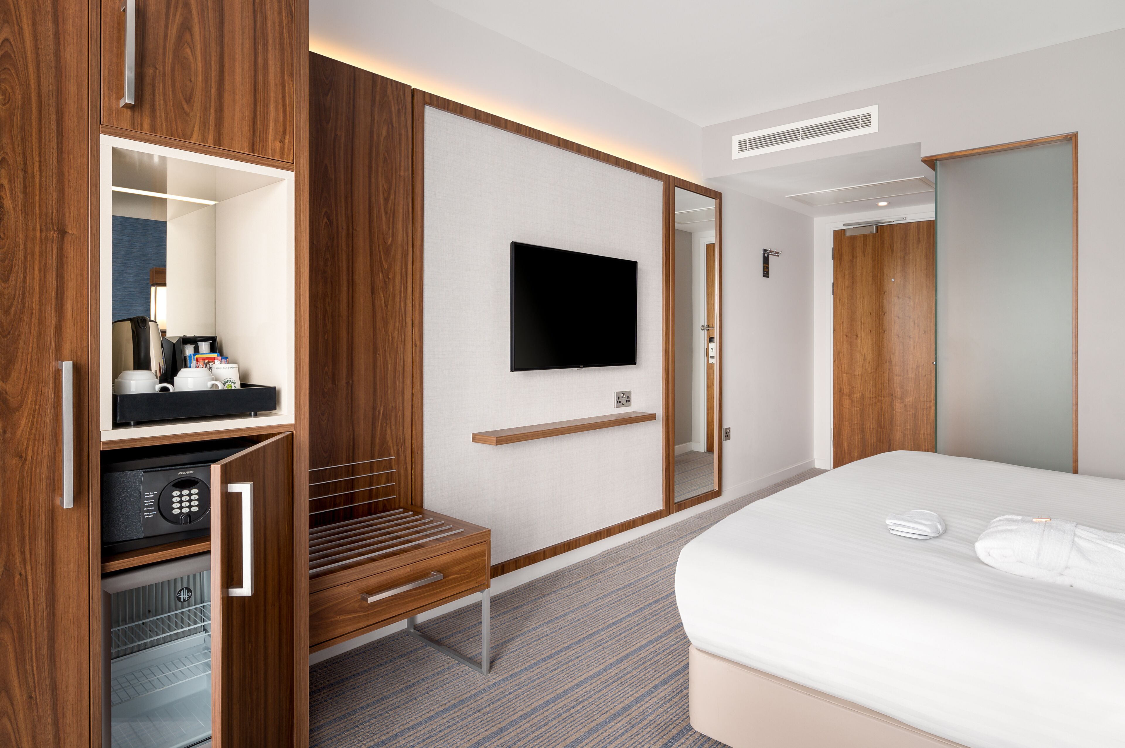 Habitación de hotel con armario de madera, televisor de pantalla plana en la pared, cama con ropa de cama blanca y un escritorio pequeño con cafetera.
