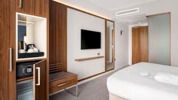 A hotel room with a wooden wardrobe, a flat-screen TV mounted on the wall, a bed with white linens, and a small desk with a coffee machine.