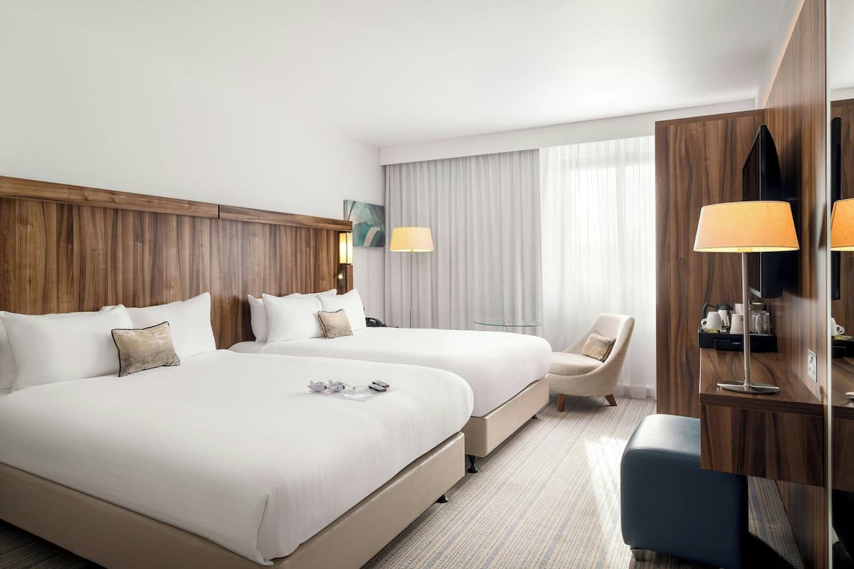 Ein Hotelzimmer mit zwei Betten, einem Schreibtisch, einem Stuhl und einem großen Fenster mit Vorhängen.