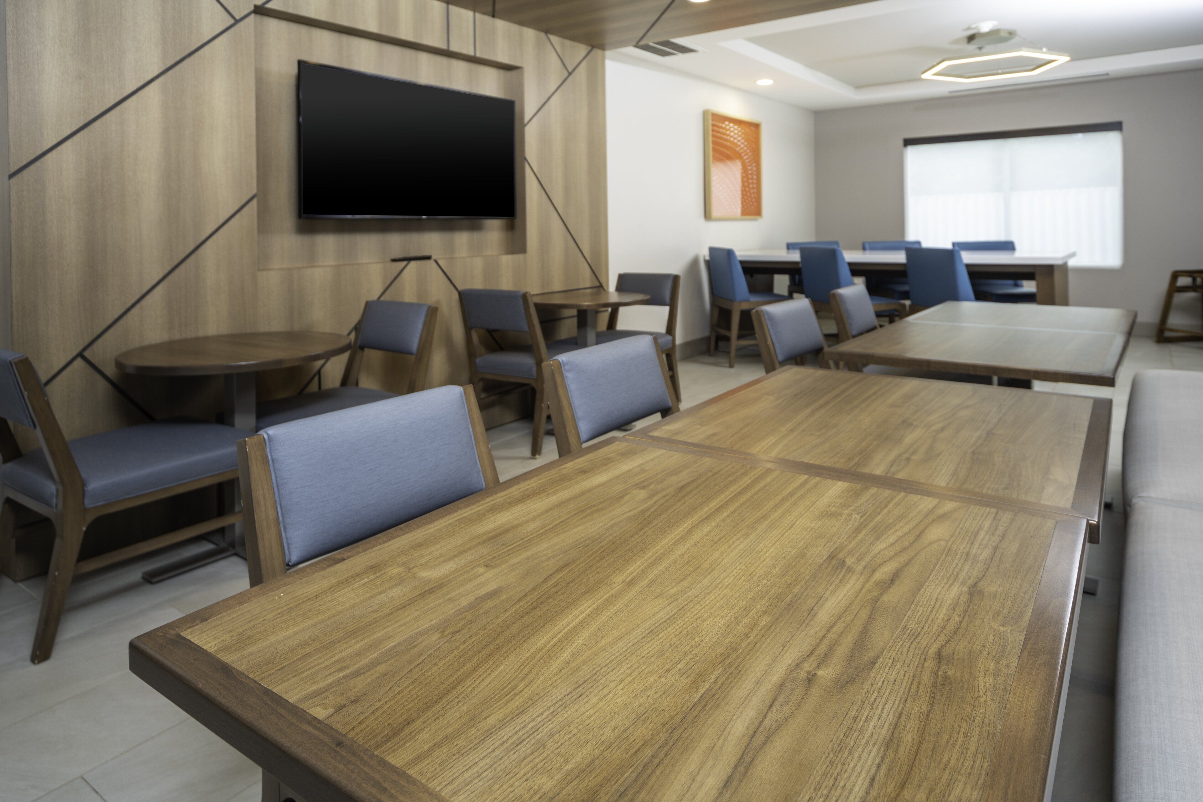 A conference room with a large wooden table, blue cushioned chairs, a flat-screen TV mounted on the wall, and a window providing natural light.
