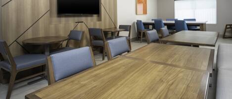 A conference room with a large wooden table, blue cushioned chairs, a flat-screen TV mounted on the wall, and a window providing natural light.
