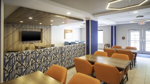 A modern dining area with wooden tables and orange chairs, a flat-screen TV mounted on the wall, and a patterned wall design. - Holiday Inn Express Hotel & Suites Atlanta Johns Creek by IHG (Suwanee)