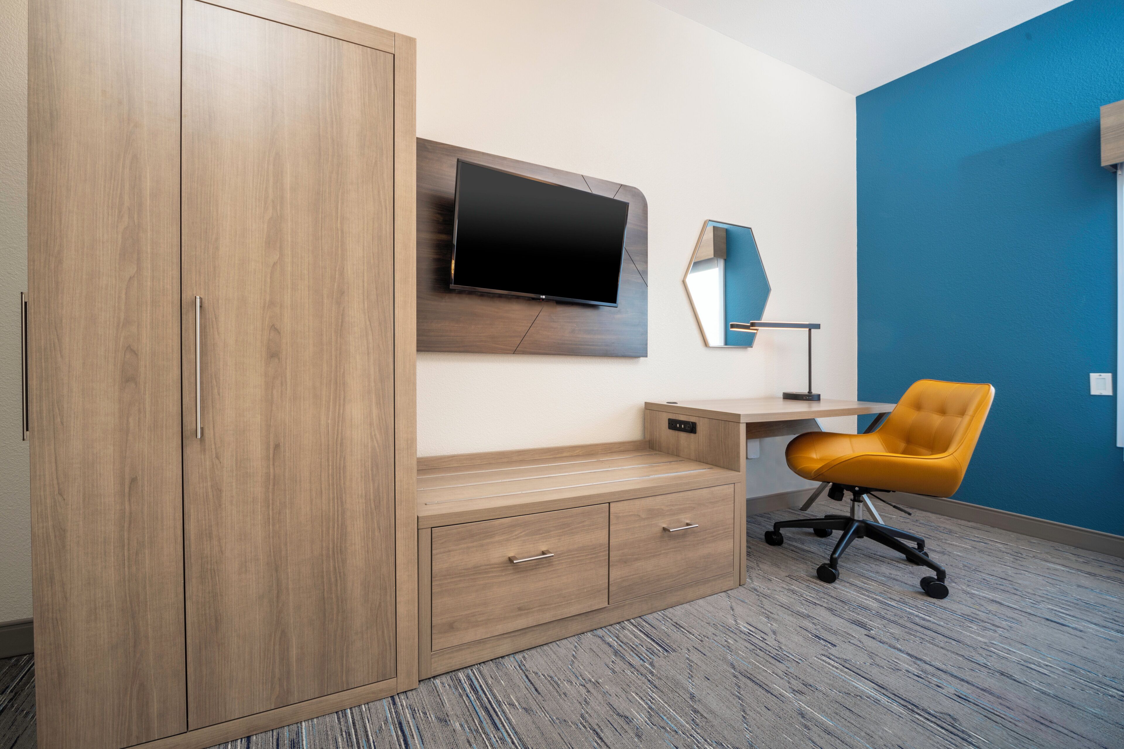 Ein Zimmer mit einem Holzkleiderschrank, einem Schreibtisch mit Flachbildfernseher, einem gelben Bürostuhl und einer blauen Akzentwand.
