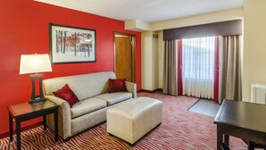 A hotel room with a red wall, a framed picture, a sofa with red cushions, a small table with a lamp, a matching ottoman, and a large window with curtains. - Holiday Inn Express & Suites Vinita by IHG (Vinita)