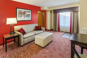 A hotel room with a red wall, a framed picture, a sofa with red cushions, a small table with a lamp, a matching ottoman, and a large window with curtains.