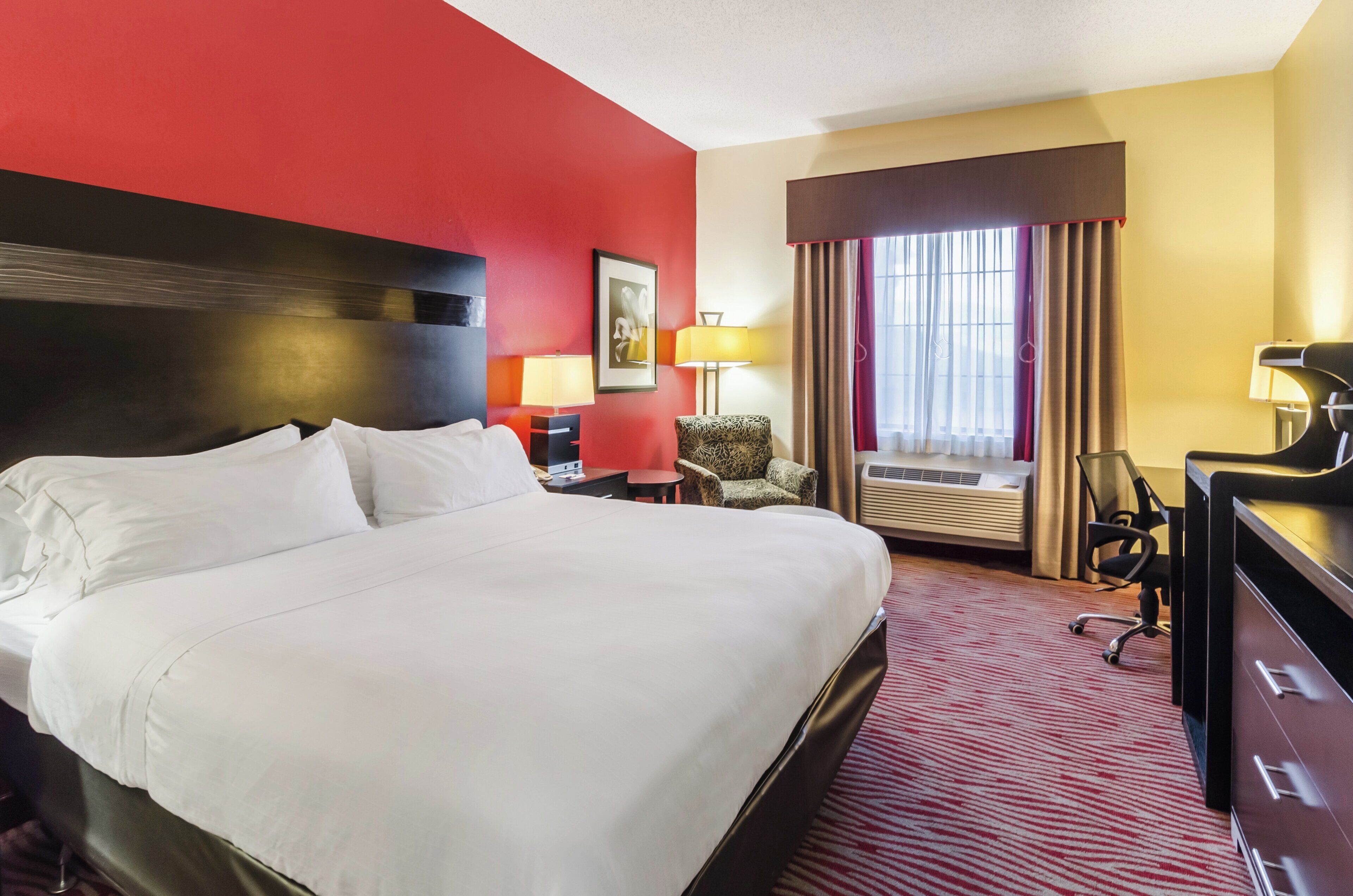 A hotel room with a large bed, a desk, a chair, a lamp, and an air conditioning unit.
