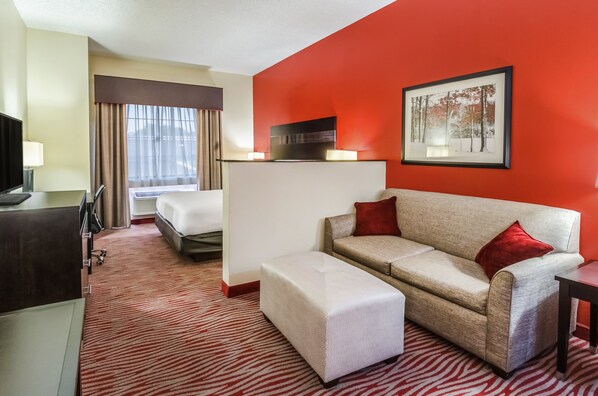 Habitación de hotel con una pared de acento rojo, un sofá beige con cojines rojos, un reposapiés blanco, un escritorio con lámpara, un televisor y un cuadro en la pared.