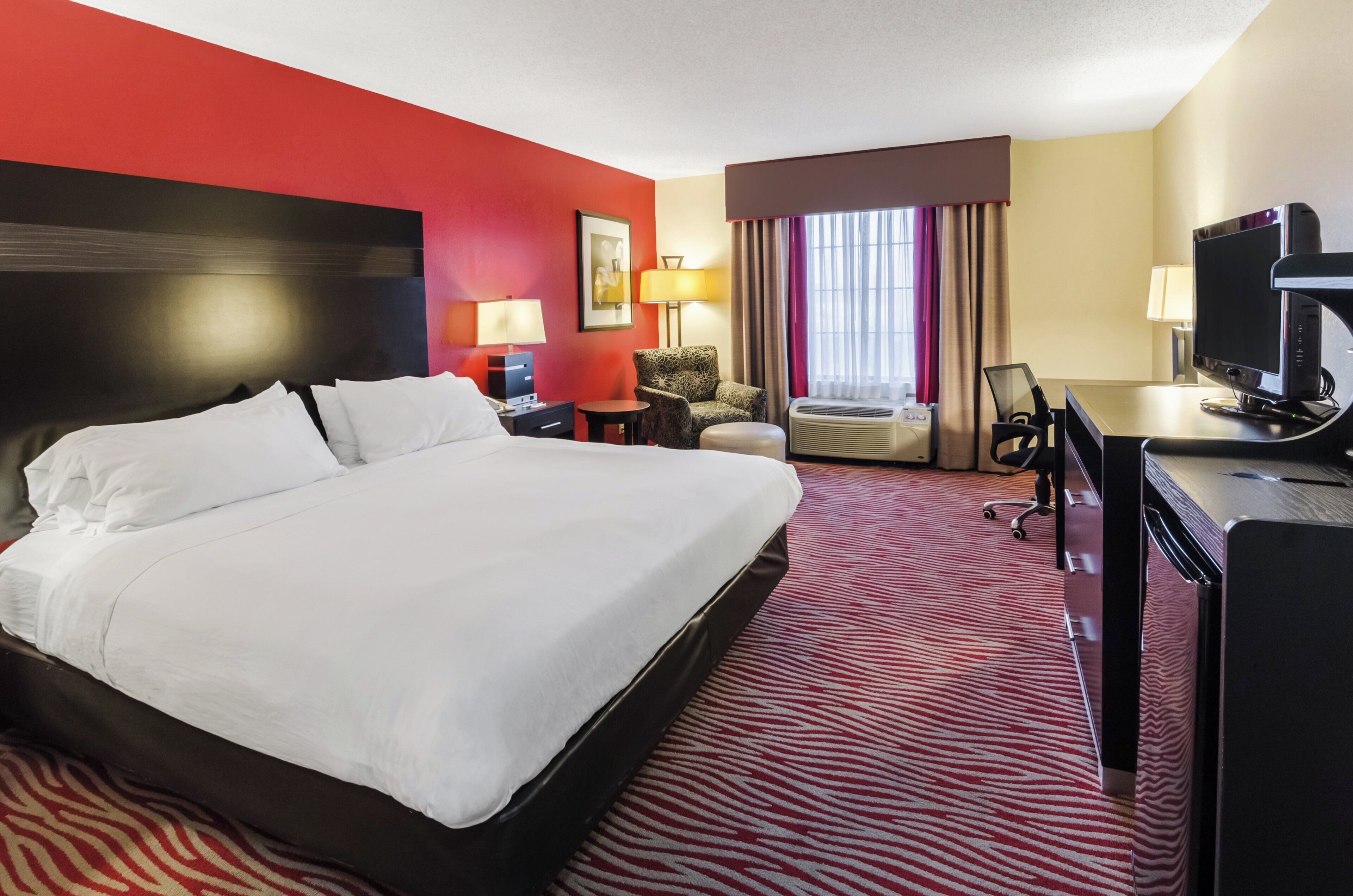 A hotel room with a large bed, a desk with a computer, a chair, a sofa, and a window with curtains.