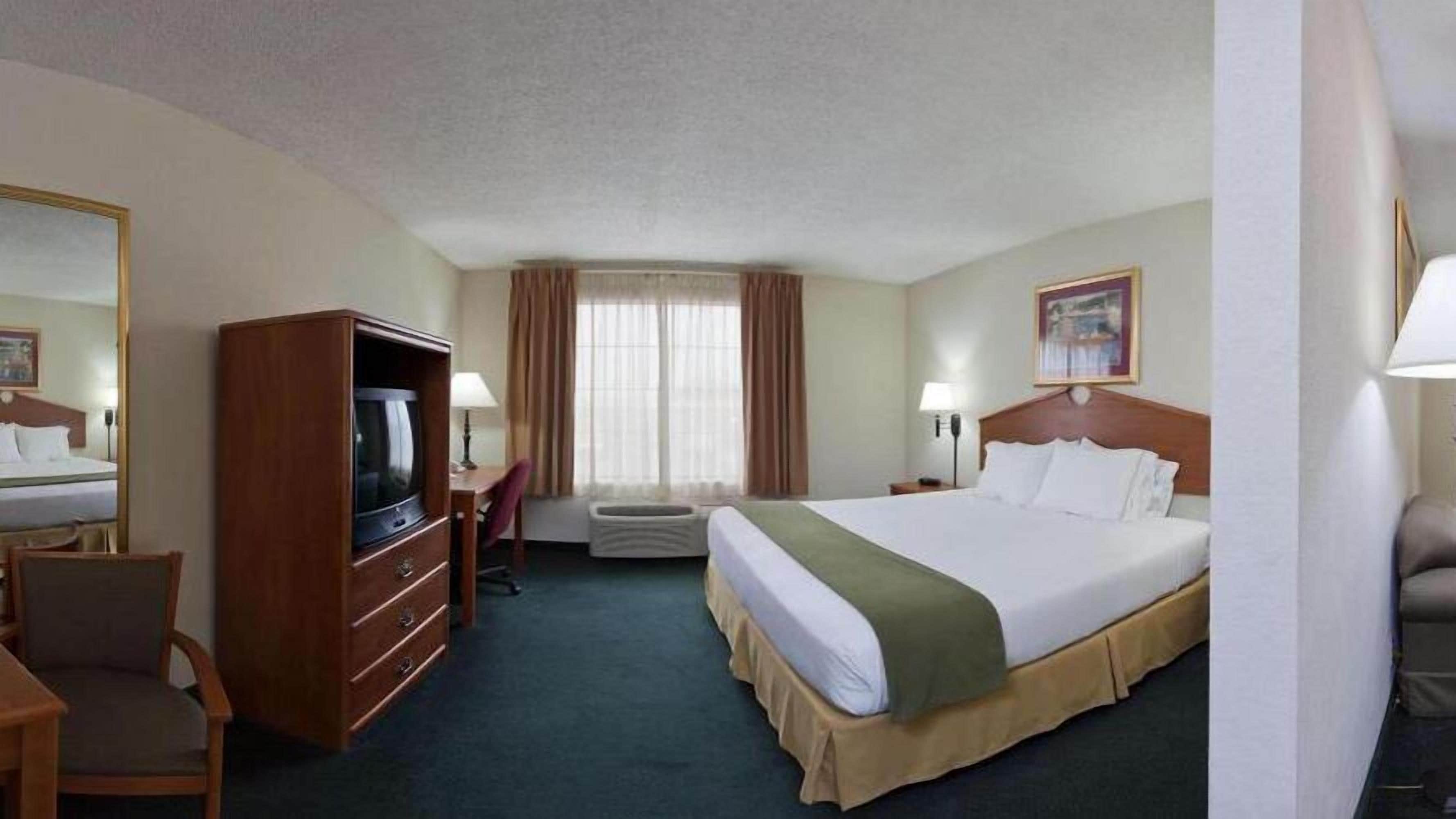 A hotel room with a large bed, a desk with a chair, a television, an air conditioning unit, and a window with curtains.
