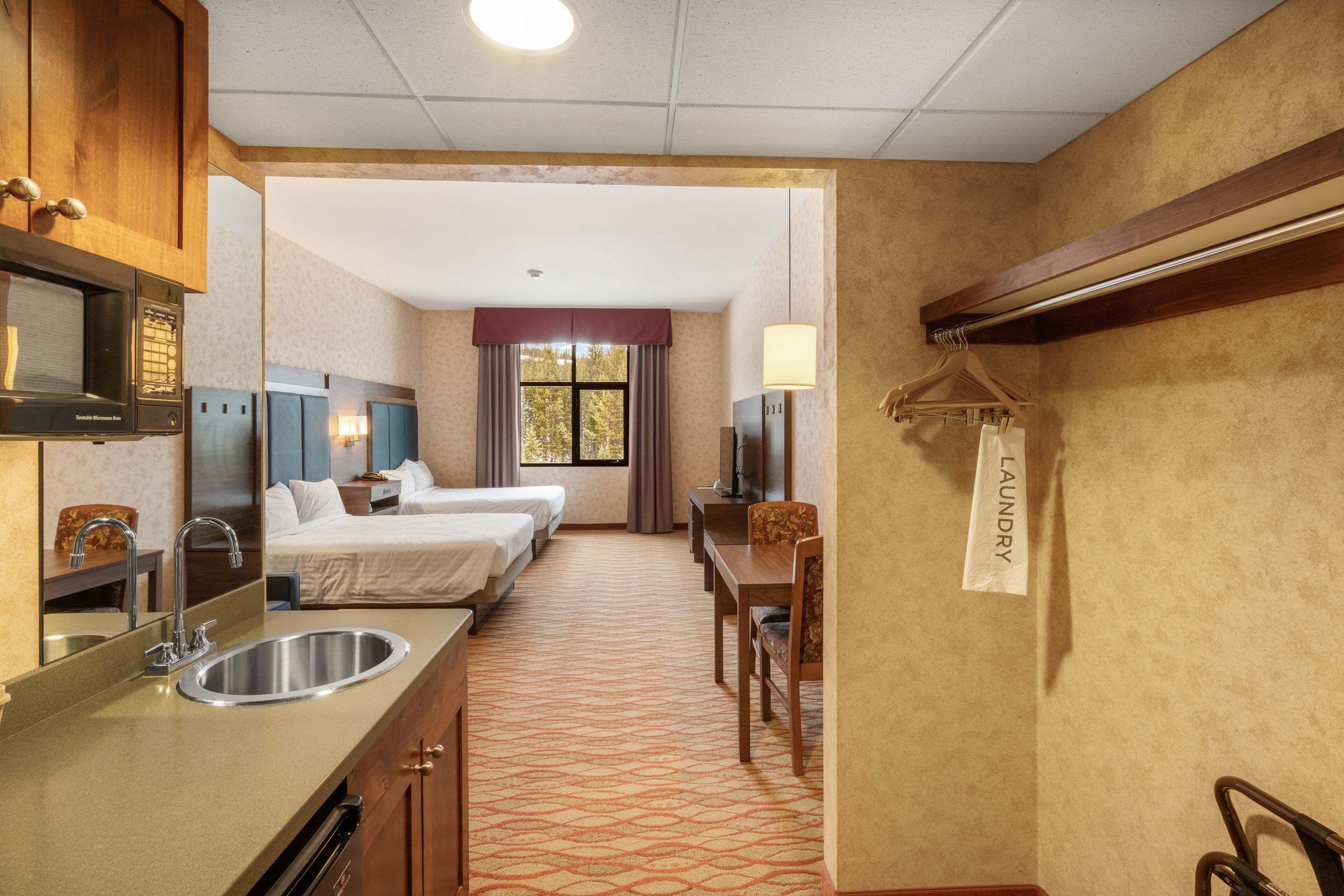 Quarto de hotel com cozinha compacta, duas camas de solteiro, escrivaninha, cadeira, televisão e janela com cortinas.