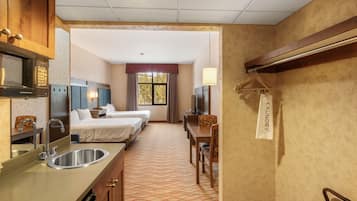 Quarto de hotel com cozinha compacta, duas camas de solteiro, escrivaninha, cadeira, televisão e janela com cortinas.