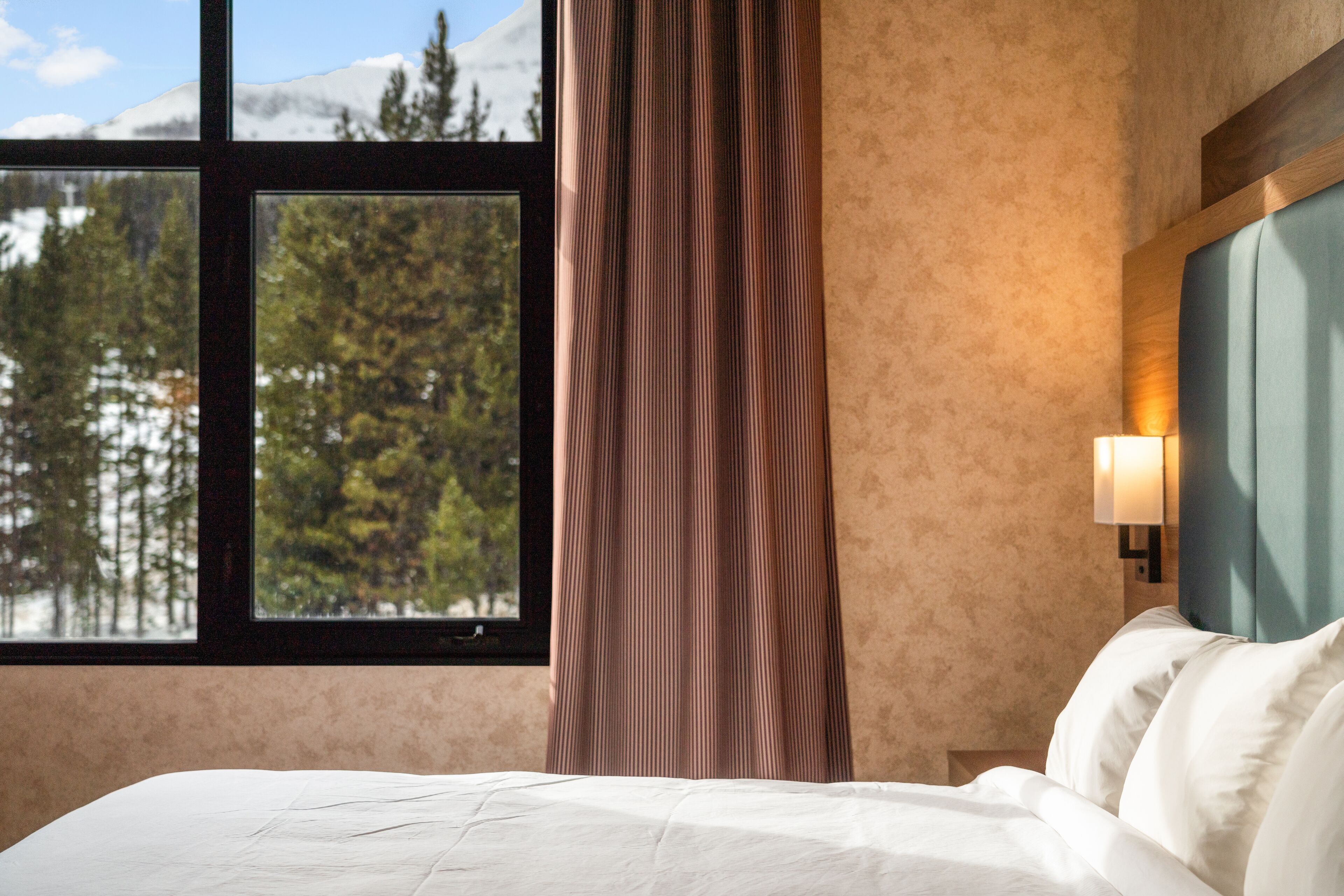 A hotel room with a bed, a window with a view of trees and mountains, and a wall-mounted lamp.