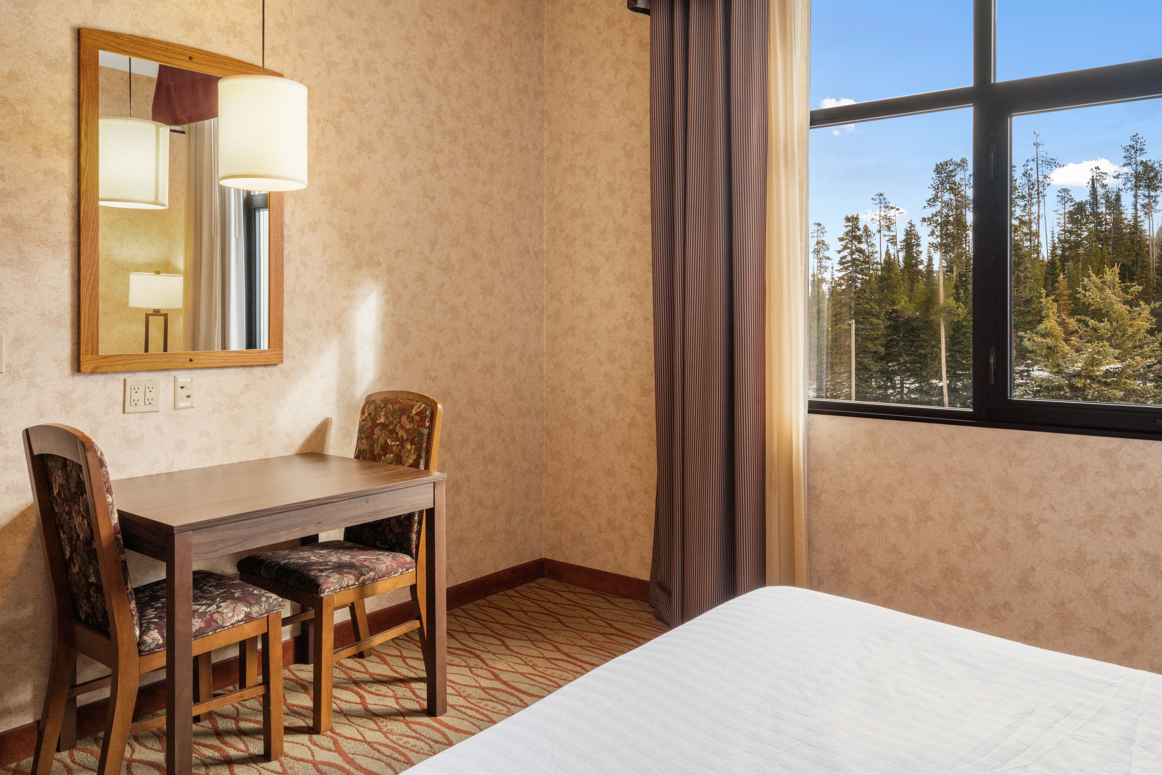 Habitación de hotel con cama, escritorio con dos sillas, espejo y ventana con vista al bosque.