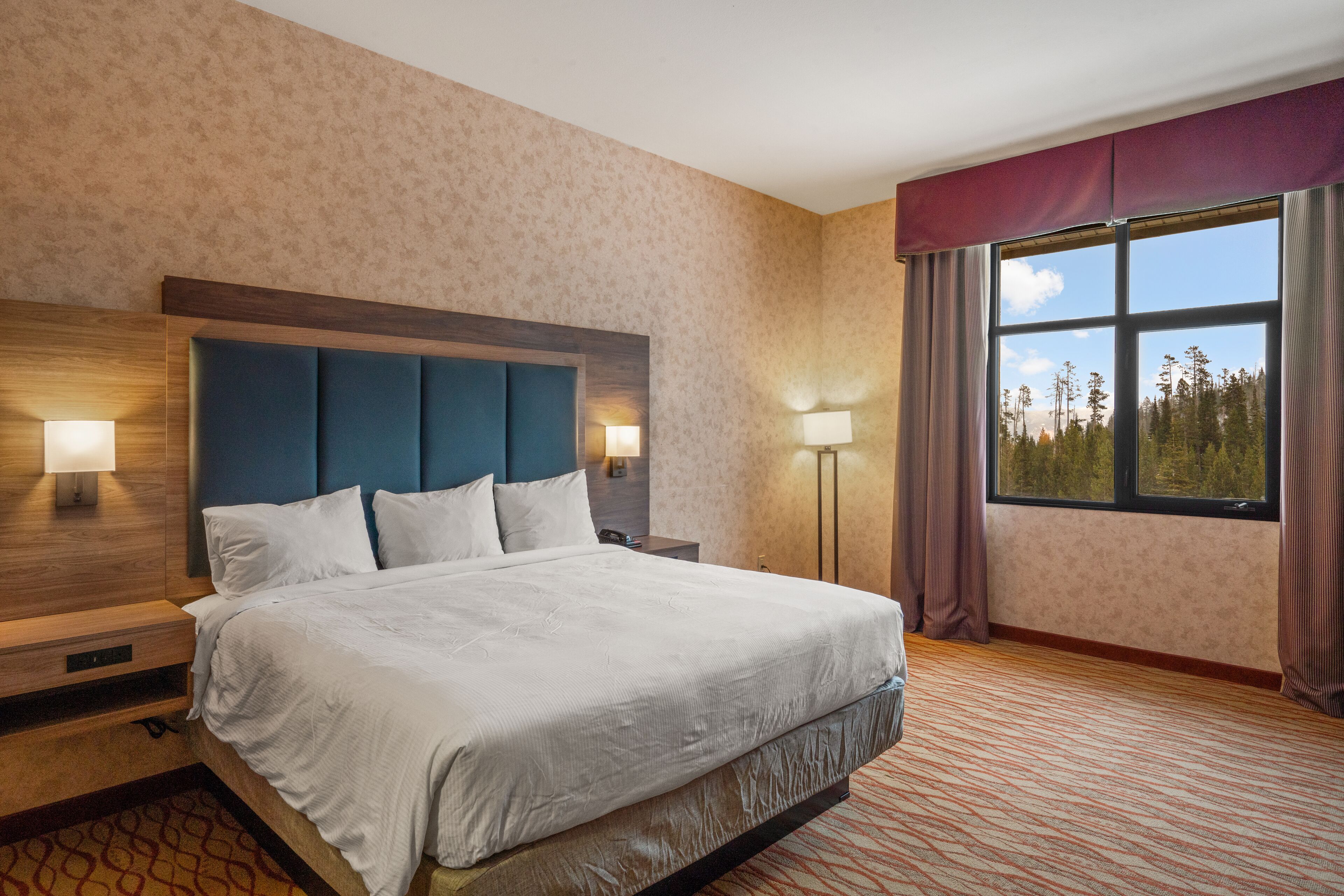 Ein Hotelzimmer mit einem großen Bett, zwei Nachttischlampen, einem Nachttisch und einem mit Vorhängen versehenen Fenster.