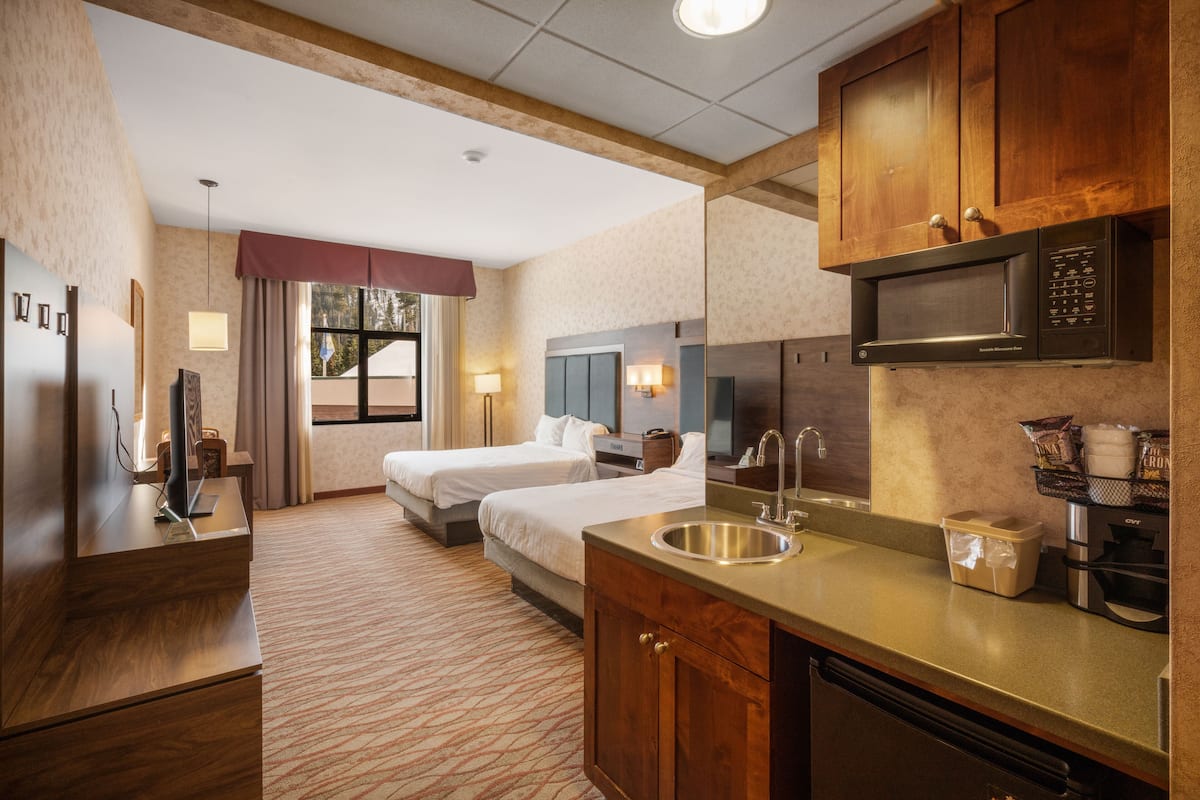 A hotel room with a kitchenette, two beds, a large window with curtains, a wooden cabinet, and a built-in microwave.